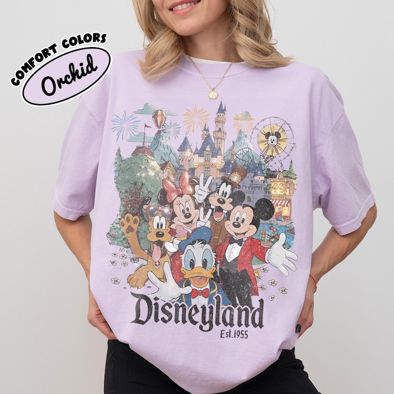 May include: Orchid Comfort Colors t-shirt featuring a Disneyland graphic with Mickey Mouse, Minnie Mouse, Donald Duck, Goofy, and other characters. The graphic includes a castle, fireworks, and the text "Disneyland Est. 1955."