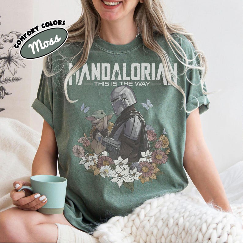 May include: Moss green t-shirt featuring a graphic of The Mandalorian and Grogu surrounded by flowers and butterflies. The text "MANDALORIAN - THIS IS THE WAY" is printed across the top. The shirt is a comfort colors style.