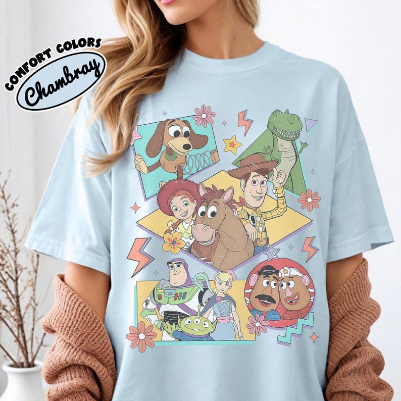 Puede incluir: Camiseta azul claro con un gr&aacute;fico colorido de personajes de la pel&iacute;cula Toy Story. El dise&ntilde;o incluye im&aacute;genes de Woody, Buzz Lightyear y otros personajes. La camiseta tiene las palabras "COMFORT COLORS Chambray" en un dise&ntilde;o circular.