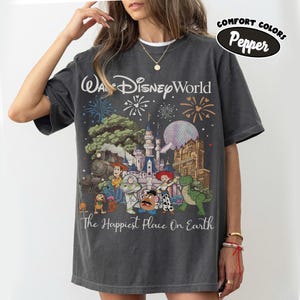 May include: Dark gray t-shirt featuring a colorful graphic of Disney World characters and fireworks. The text "Wa Disney World" and "The Happiest Place On Earth" are printed on the front. The shirt is labeled "Comfort Colors Pepper".