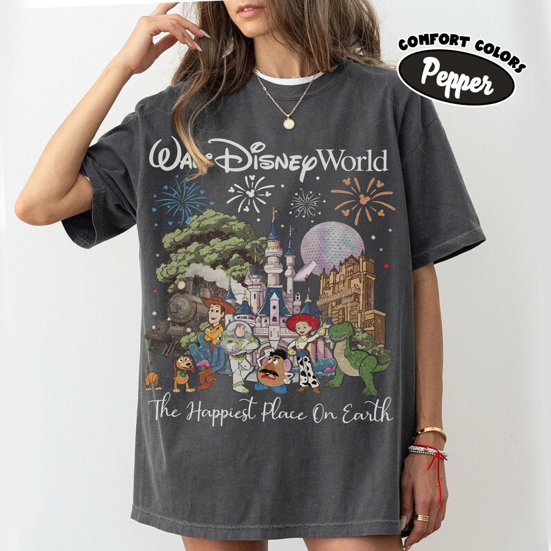 May include: Dark gray t-shirt featuring a colorful graphic of Disney World characters and fireworks. The text "Wa Disney World" and "The Happiest Place On Earth" are printed on the front. The shirt is labeled "Comfort Colors Pepper".