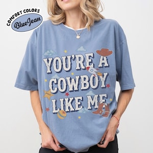 May include: A blue short-sleeve t-shirt featuring the text "YOU'RE A COWBOY LIKE ME" in white letters. The graphic includes a brown cowboy hat, a rocket ship, a yellow sheriff star, a ball, and cowboy boots, surrounded by small red stars and clouds.