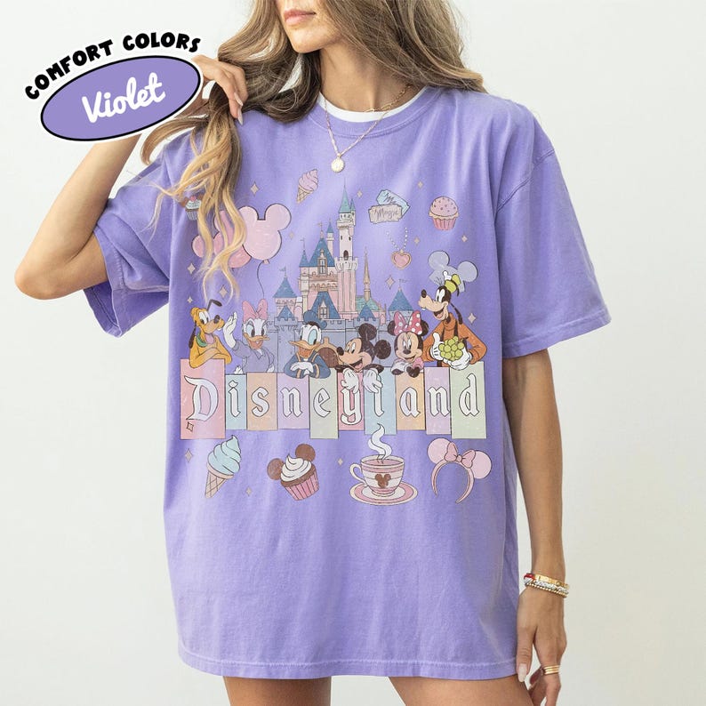 May include: A violet short-sleeve t-shirt featuring a graphic print of a castle, Mickey Mouse, Minnie Mouse, Donald Duck, Daisy Duck, Goofy, and Pluto. The text "Disneyland" is printed across the center, accompanied by icons of cupcakes, ice cream, and mouse ears.