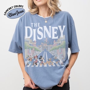 Może przedstawiać: Niebieska koszulka z kr&oacute;tkim rękawem z napisem "THE DISNEY" z przodu. Grafika przedstawia postacie Disneya przechodzące przez ulicę z zamkiem w tle. Koszulka ma logo "Comfort Colors Blue Jean".