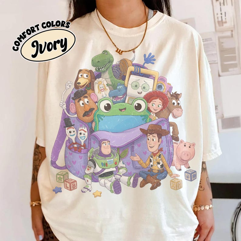 Pode incluir: Uma t-shirt cor de marfim com uma impress&atilde;o gr&aacute;fica de v&aacute;rias personagens animadas de Toy Story, incluindo um ranger espacial, um cowboy, um dinossauro verde e um porco, reunidos &agrave; volta de uma mochila roxa com uma cara de r&atilde; verde.