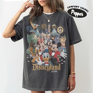 May include: Dark gray t-shirt featuring a Disneyland graphic with Mickey Mouse, Minnie Mouse, Donald Duck, and Goofy. The graphic includes a castle, fireworks, and the text "Disneyland Est.1955". The shirt has short sleeves and a crew neck.
