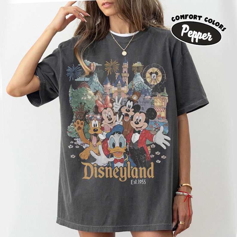 May include: Dark gray t-shirt featuring a Disneyland graphic with Mickey Mouse, Minnie Mouse, Donald Duck, and Goofy. The graphic includes a castle, fireworks, and the text "Disneyland Est.1955". The shirt has short sleeves and a crew neck.