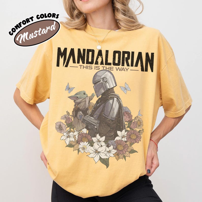 May include: Mustard-colored t-shirt featuring the text "MANDALORIAN" above "THIS IS THE WAY." The graphic depicts the Mandalorian and Grogu surrounded by flowers and butterflies. The shirt is a comfort color.