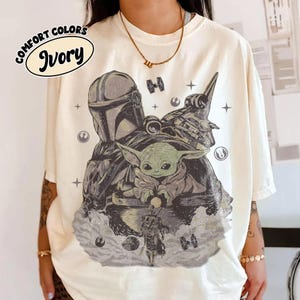 Puede incluir: Camiseta marfil Comfort Colors con un gr&aacute;fico de The Mandalorian, Grogu y una nave espacial. El dise&ntilde;o incluye el texto "Comfort Colors" e "Ivory" en un dise&ntilde;o circular. La obra de arte es en tonos grises, verdes y blancos.