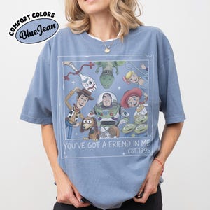 May include: Blue t-shirt with a graphic featuring characters from the animated film "Toy Story." The design includes Woody, Buzz Lightyear, and other characters, with the text "YOU'VE GOT A FRIEND IN ME EST.1995" in a white frame. The shirt has the words "COMFORT COLORS Blue Jean" in a circle.