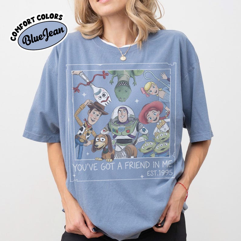 May include: Blue t-shirt with a graphic featuring characters from the animated film "Toy Story." The design includes Woody, Buzz Lightyear, and other characters, with the text "YOU'VE GOT A FRIEND IN ME EST.1995" in a white frame. The shirt has the words "COMFORT COLORS Blue Jean" in a circle.