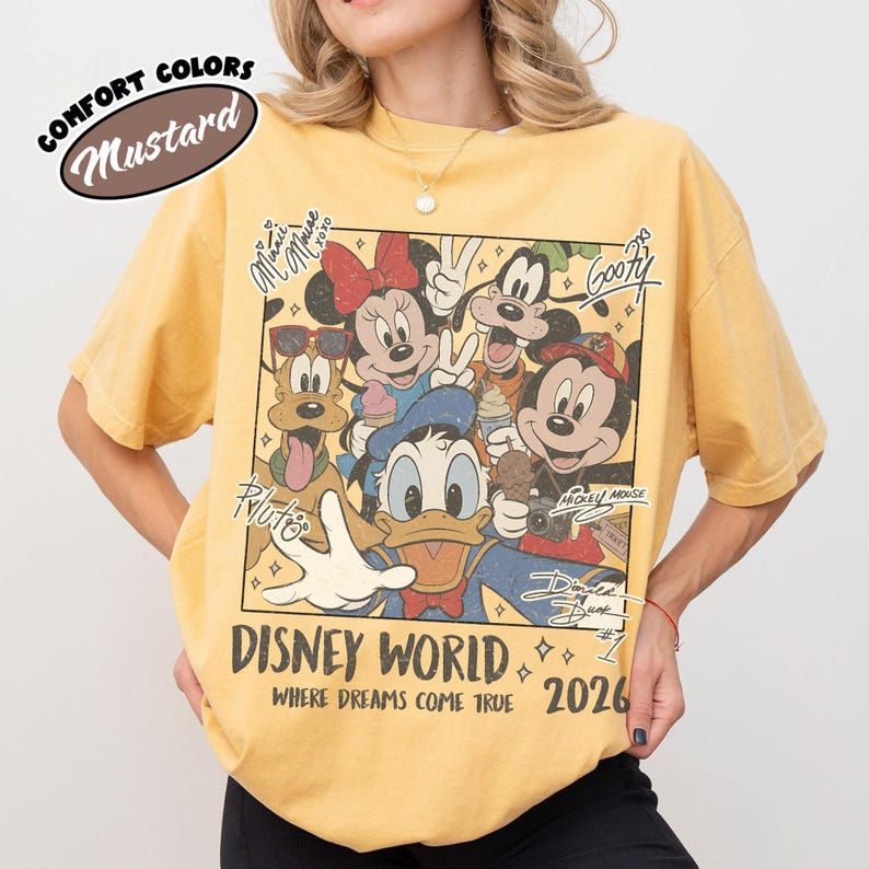 May include: Mustard-colored t-shirt featuring a graphic of Disney characters, including Mickey Mouse, Minnie Mouse, Donald Duck, and Goofy. The shirt has the text "DISNEY WORLD" and "2026" printed on it. The shirt is a Comfort Colors brand.