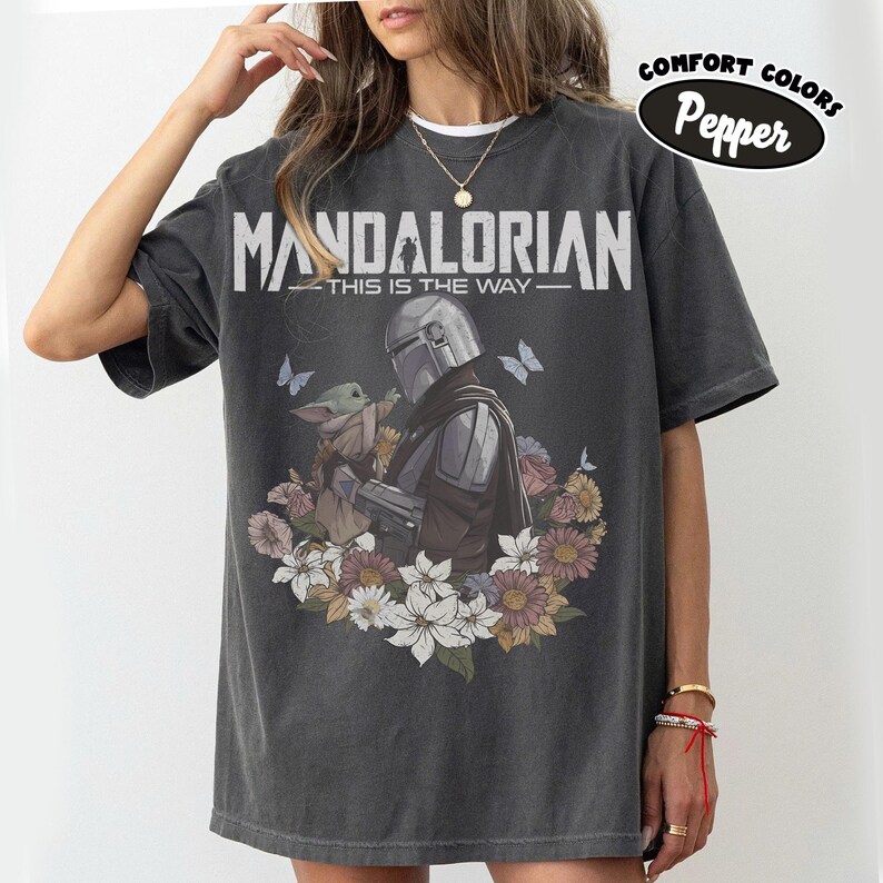 May include: Dark gray t-shirt featuring a graphic of The Mandalorian and Grogu surrounded by flowers and butterflies. The text "MANDALORIAN - THIS IS THE WAY" is printed above the image. The shirt has a Comfort Colors Pepper label.