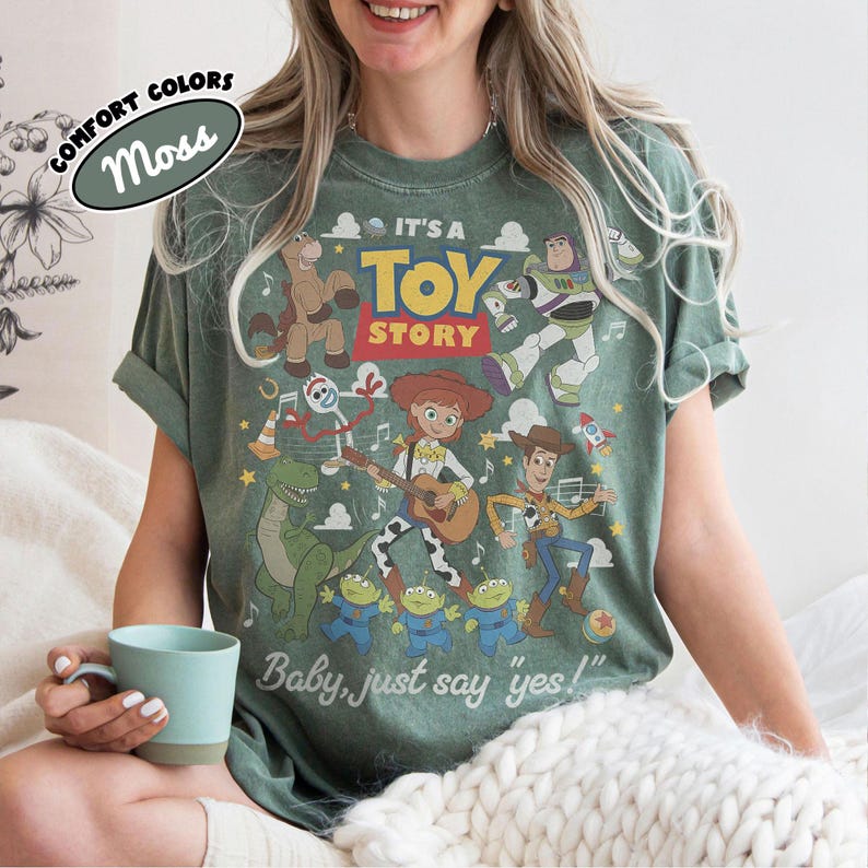 May include: A moss green short-sleeve t-shirt featuring a graphic print of Toy Story characters including Woody, Jessie, Buzz Lightyear, Rex, and Forky. The text reads IT'S A TOY STORY and Baby, just say yes! with musical notes and stars.