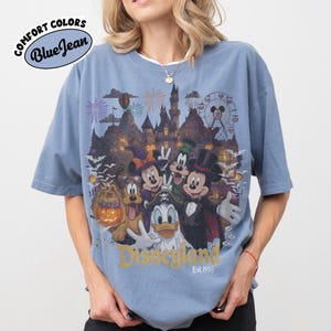 May include: A blue short-sleeve t-shirt featuring a graphic of Disney characters in Halloween costumes, including Mickey Mouse, Minnie Mouse, Goofy, Pluto, and Donald Duck, with a castle, fireworks, and the text Disneyland Est. 1955.