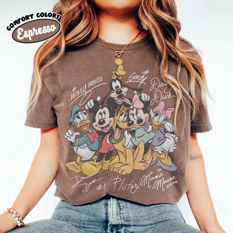 May include: Espresso-colored t-shirt featuring a graphic of Disney characters Mickey Mouse, Donald Duck, Goofy, Pluto, Minnie Mouse, and Daisy Duck. The shirt has the text "Mickey Mouse" and character signatures. The shirt is a comfort colors tee.