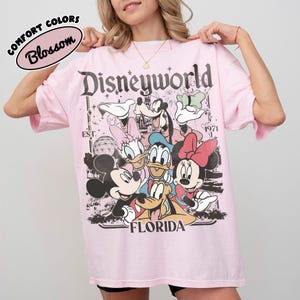 May include: A light pink t-shirt featuring a graphic of Disney characters, including Mickey and Minnie Mouse, Donald and Daisy Duck, and Goofy. The text "Disneyworld Florida" is printed on the front. The shirt also has the text "Comfort Colors Blossom".