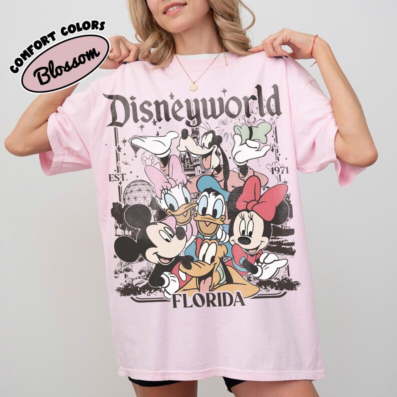 May include: A light pink t-shirt featuring a graphic of Disney characters, including Mickey and Minnie Mouse, Donald and Daisy Duck, and Goofy. The text "Disneyworld Florida" is printed on the front. The shirt also has the text "Comfort Colors Blossom".