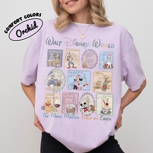 May include: A light purple t-shirt with a Walt Disney World design. The shirt features various Disney characters and country stamps. The text "COMFORT COLORS Orchid" is printed on the top left.