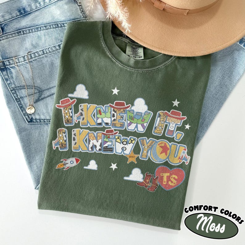 May include: An olive green t-shirt featuring a graphic print with the text I KNEW IT, I KNEW YOU. The design includes colorful patterns inspired by cowboy and space themes, small white stars, cloud shapes, a rocket, and a red heart with the letters TS.