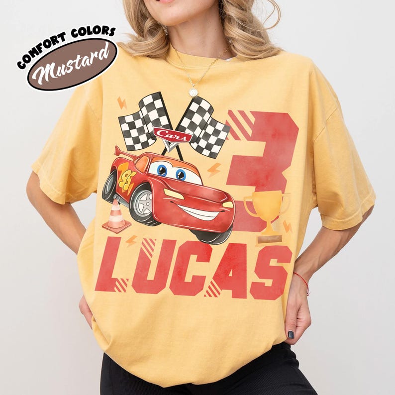 Puede incluir: Camiseta color mostaza con un coche de carreras rojo de dibujos animados, el n&uacute;mero 95, banderas a cuadros y el nombre "LUCAS". El n&uacute;mero "3" y un trofeo dorado tambi&eacute;n se muestran.
