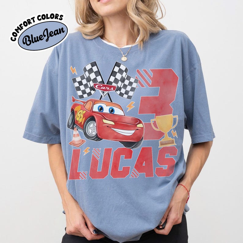 Puede incluir: Camiseta azul con un gr&aacute;fico de un coche de carreras rojo, banderas a cuadros y el n&uacute;mero 3. El nombre "LUCAS" est&aacute; impreso en letras rojas debajo del coche. La camiseta tiene las palabras "Comfort Colors" y "Blue Jean" impresas.