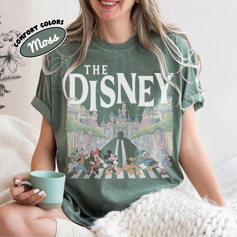 Może przedstawiać: Koszulka w kolorze mchu z białym napisem "THE DISNEY" nad kolorową ilustracją postaci Disneya przechodzących przez ulicę przed zamkiem. Koszulka ma podwinięte rękawy, a na etykiecie widnieją napisy "COMFORT COLORS" i "Moss".