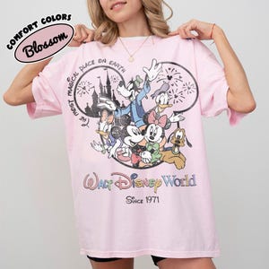 May include: Pink t-shirt featuring a graphic of Disney characters including Mickey Mouse, Minnie Mouse, Donald Duck, and Goofy. The shirt has the text "The Most Magical Place on Earth" and "Walt Disney World Since 1971". The shirt is labeled "Comfort Colors Blossom".