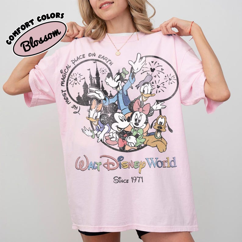 May include: Pink t-shirt featuring a graphic of Disney characters including Mickey Mouse, Minnie Mouse, Donald Duck, and Goofy. The shirt has the text "The Most Magical Place on Earth" and "Walt Disney World Since 1971". The shirt is labeled "Comfort Colors Blossom".