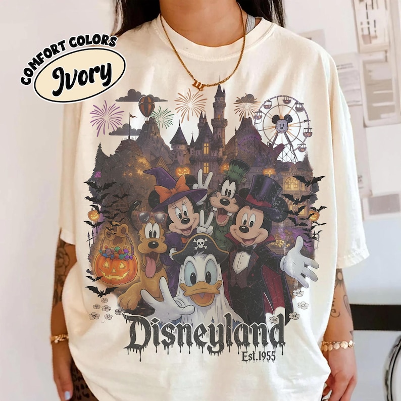 May include: An ivory t-shirt featuring a graphic of Disney characters in Halloween costumes, including Mickey Mouse as a vampire, Minnie Mouse as a witch, and Donald Duck as a ghost, with a castle, ferris wheel, and the text Disneyland Est. 1955.