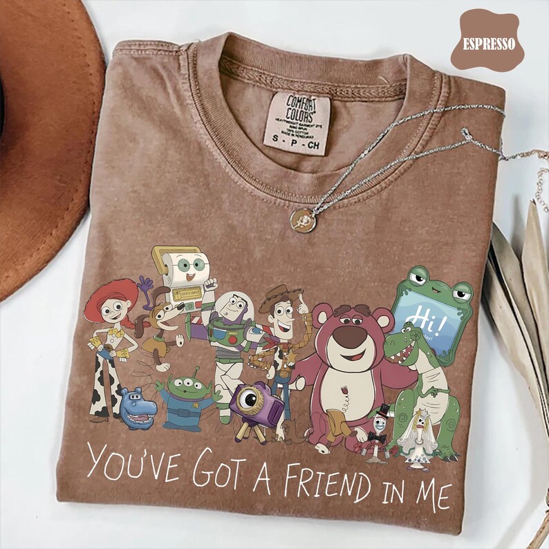 May include: A brown short-sleeved t-shirt featuring a graphic print of various animated characters from the Toy Story franchise. The text "YOU'VE GOT A FRIEND IN ME" is printed in white at the bottom. The shirt has a Comfort Colors brand label.