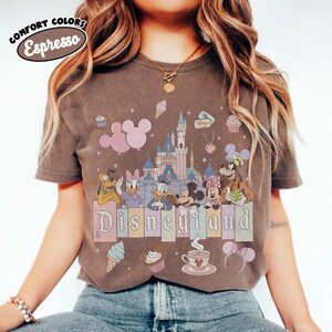 May include: A dark brown short-sleeve t-shirt featuring a graphic print of a castle, Mickey Mouse, Minnie Mouse, Donald Duck, Daisy Duck, Goofy, and Pluto. The text "Disneyland" is printed in the center, with "COMFORT COLORS Espresso" at the top.