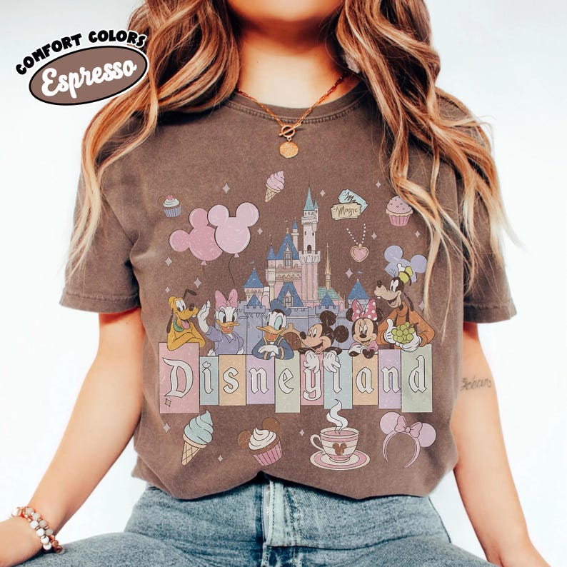 May include: A dark brown short-sleeve t-shirt featuring a graphic print of a castle, Mickey Mouse, Minnie Mouse, Donald Duck, Daisy Duck, Goofy, and Pluto. The text "Disneyland" is printed in the center, with "COMFORT COLORS Espresso" at the top.