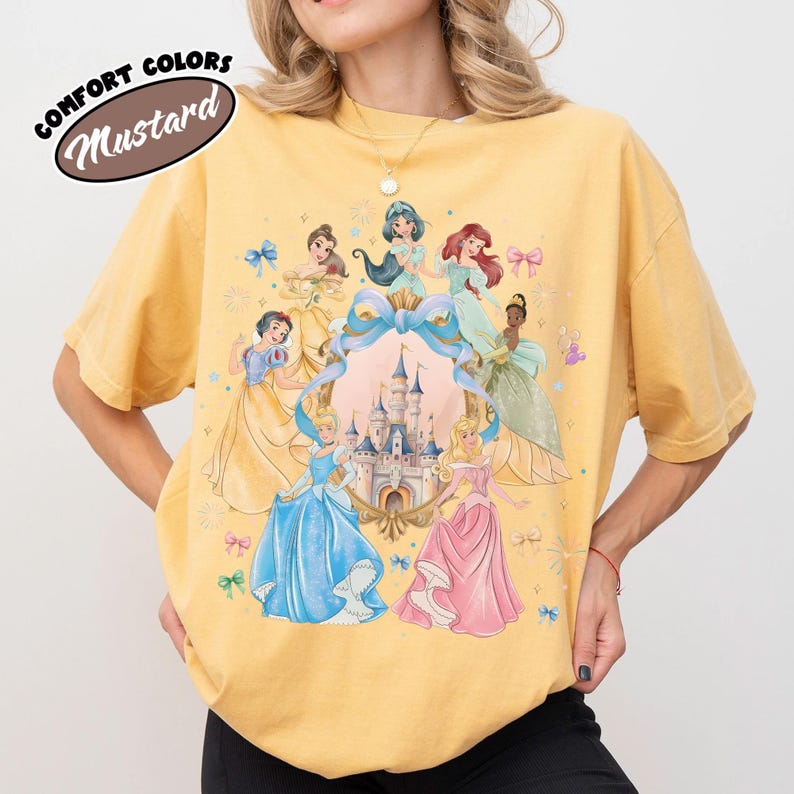 May include: Mustard-colored t-shirt featuring a graphic of several animated princesses surrounding a castle. The princesses are in various gowns, with a large blue bow and a castle in the center. The text "COMFORT COLORS" and "Mustard" are in a brown oval.