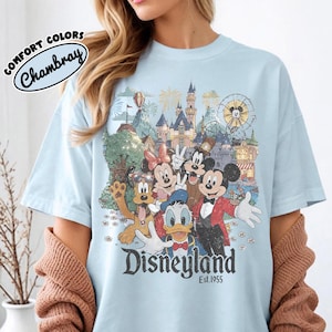 May include: Light blue t-shirt featuring a vintage Disneyland graphic with Mickey Mouse, Minnie Mouse, Donald Duck, and Pluto. The shirt has the text "Disneyland Est. 1955" and "Comfort Colors Chambray" on the upper left.