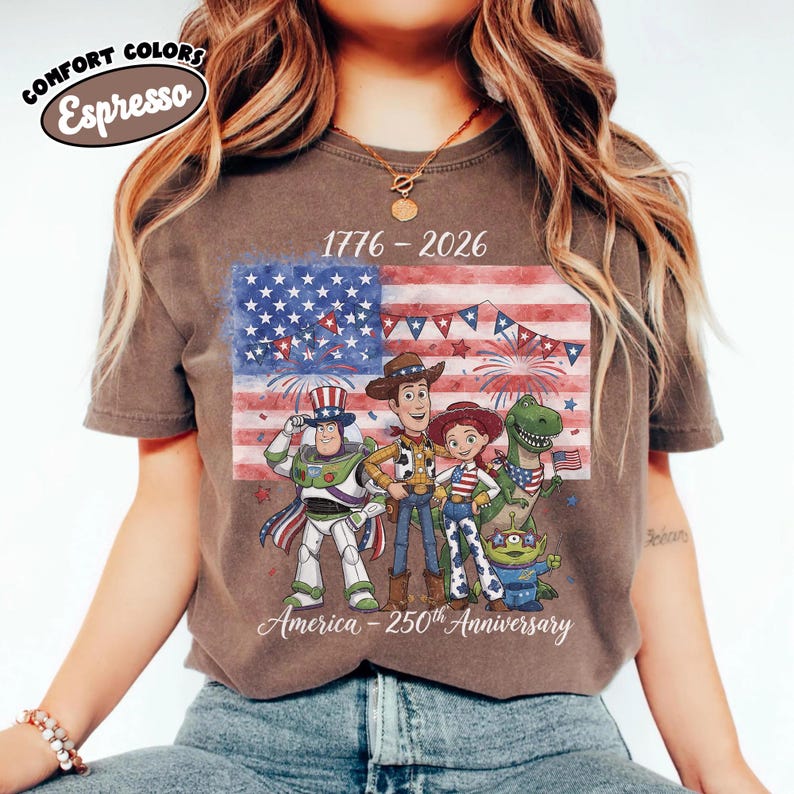 Może przedstawiać: T-shirt w kolorze espresso z grafiką amerykańskiej flagi, postaciami z kresk&oacute;wek oraz tekstem "1776-2026" i "America - 250th Anniversary". Koszulka ma okrągły dekolt i kr&oacute;tkie rękawy.