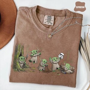 May include: An espresso-colored short-sleeve t-shirt featuring a graphic print of five green creatures in robes. One holds a spear, one a slingshot, one a net, one a white helmet, and one is sleeping. The shirt has a Comfort Colors brand label.