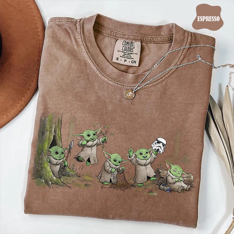 May include: An espresso-colored short-sleeve t-shirt featuring a graphic print of five green creatures in robes. One holds a spear, one a slingshot, one a net, one a white helmet, and one is sleeping. The shirt has a Comfort Colors brand label.