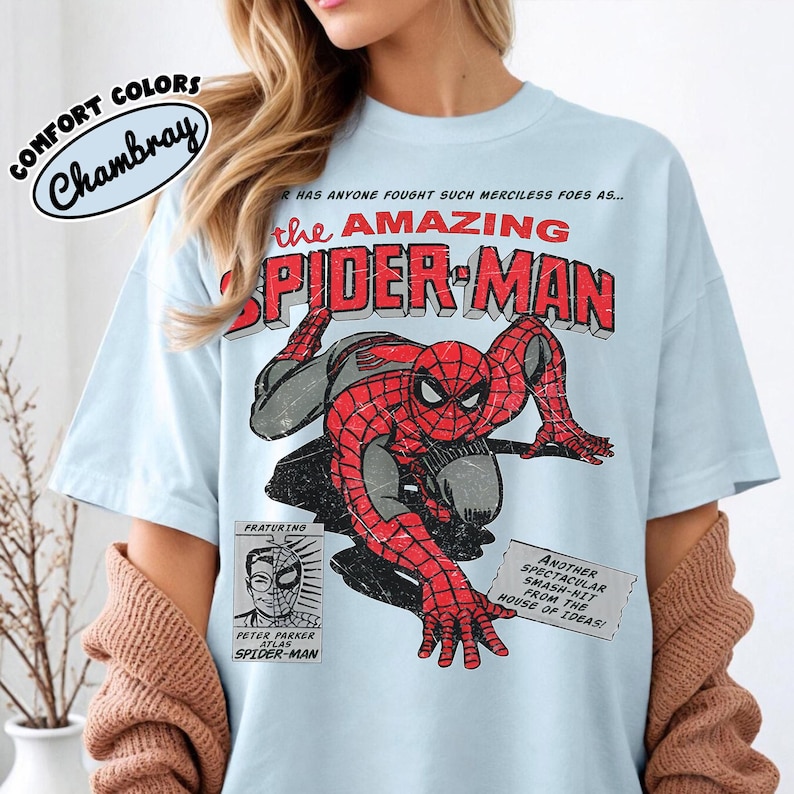 May include: A light blue t-shirt featuring a graphic of Spider-Man in a crouching pose. The text reads: HAS ANYONE FOUGHT SUCH MERCILESS FOES AS... THE AMAZING SPIDER-MAN, PRATURING PETER PARKER ATLAS SPIDER-MAN, ANOTHER SPECTACULAR SMASH-HIT FROM THE HOUSE OF IDEAS!