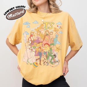 May include: A mustard yellow t-shirt featuring a graphic print of various animated characters in a collage style. The design includes the text "TS" at the top, a rocket, clouds, a star-shaped badge, and a camera. The shirt has short sleeves.