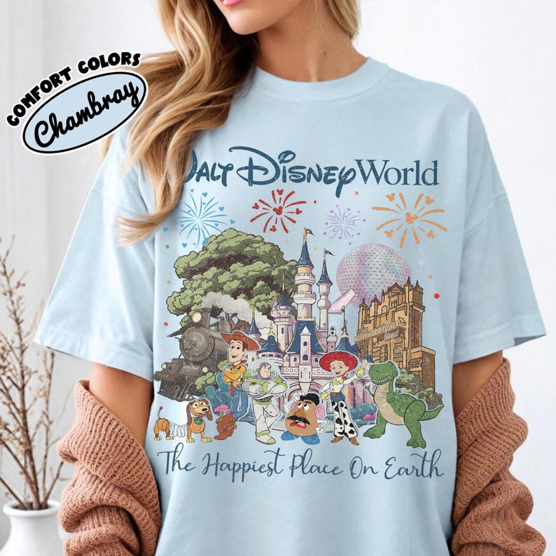 May include: Light blue t-shirt with a Walt Disney World graphic featuring various characters and attractions. The text "The Happiest Place On Earth" is printed below the graphic. The shirt has the words "Comfort Colors" and "Chambray" printed on the upper left.