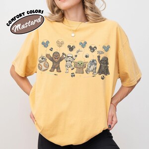 K&ouml;nnte beinhalten: Ein senfgelbes Kurzarm-T-Shirt mit einem Grafikdruck verschiedener Star-Wars-Figuren, darunter Chewbacca, ein Sturmtruppler, Grogu, R2-D2 und Darth Vader, die Luftballons halten. Der Text COMFORT COLORS MUSTARD ist in der Ecke aufgedruckt.