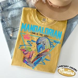 Może przedstawiać: Musztardowa koszulka z grafiką The Mandalorian i Grogu, z napisem "MANDALORIAN - THIS IS THE WAY" w kolorze niebieskim. Koszulka jest złożona na niebieskich dżinsach, z beżowym kapeluszem i srebrnym naszyjnikiem.