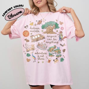 Puede incluir: Camiseta rosa claro con un dise&ntilde;o gr&aacute;fico colorido que presenta personajes de dibujos animados, un &aacute;rbol y las palabras "Hakuna Matata" y "Adventure is out there". La camiseta tambi&eacute;n tiene las palabras "anyone can be anything".