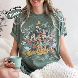 May include: Moss green t-shirt featuring a graphic of Mickey Mouse and friends. The design includes Mickey, Minnie, Donald, Daisy, Goofy, and Pluto, with their names in a vintage style. The shirt has rolled-up sleeves and the words "Comfort Colors" and "Moss" are printed on the top left.