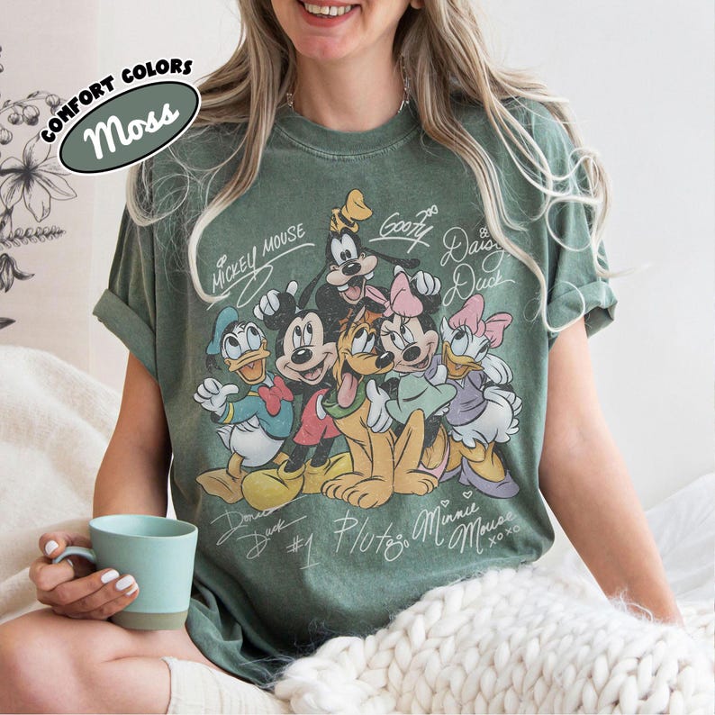 May include: Moss green t-shirt featuring a graphic of Mickey Mouse and friends. The design includes Mickey, Minnie, Donald, Daisy, Goofy, and Pluto, with their names in a vintage style. The shirt has rolled-up sleeves and the words "Comfort Colors" and "Moss" are printed on the top left.