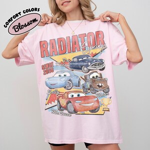May include: A light pink t-shirt featuring a graphic of characters from the movie "Cars." The design includes the words "Radiator Springs" and the names of the characters. The shirt has short sleeves and a crew neck.