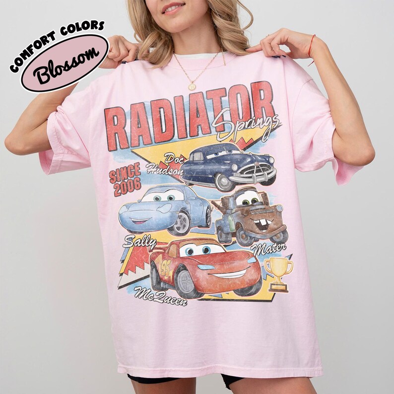 May include: A light pink t-shirt featuring a graphic of characters from the movie "Cars." The design includes the words "Radiator Springs" and the names of the characters. The shirt has short sleeves and a crew neck.