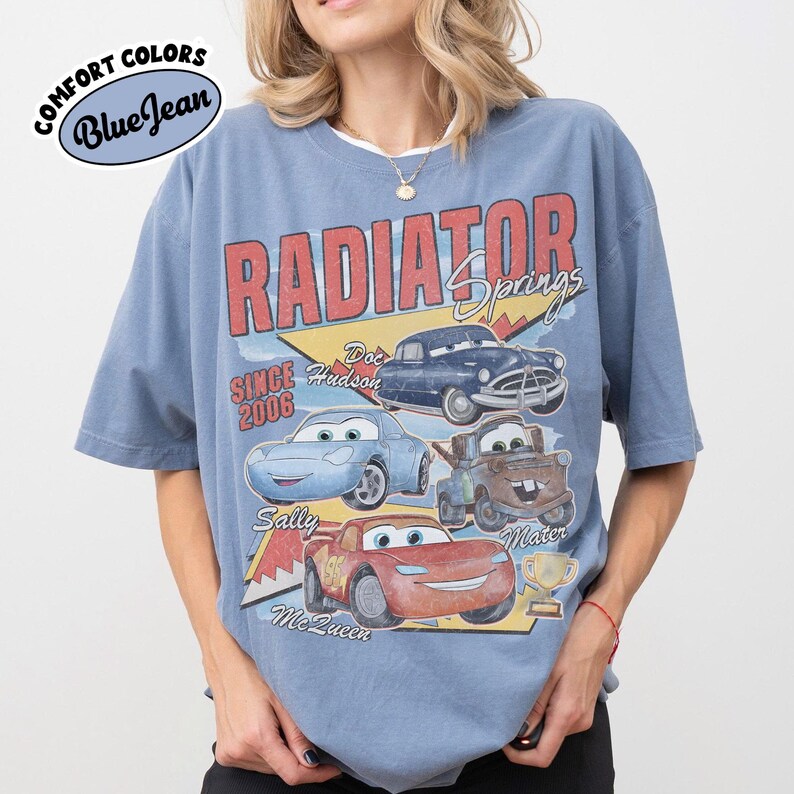 May include: Blue t-shirt with a graphic of characters from the movie Cars, including Lightning McQueen, Sally, and Mater. The shirt features the text "Radiator Springs" and "Since 2006". The shirt is labeled "Comfort Colors Blue Jean".
