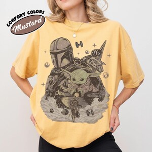 Puede incluir: Camiseta color mostaza con un gr&aacute;fico de The Mandalorian, Grogu y una nave espacial en tonos grises y marrones. El texto "COMFORT COLORS" y "Mustard" est&aacute; impreso en la esquina superior izquierda.